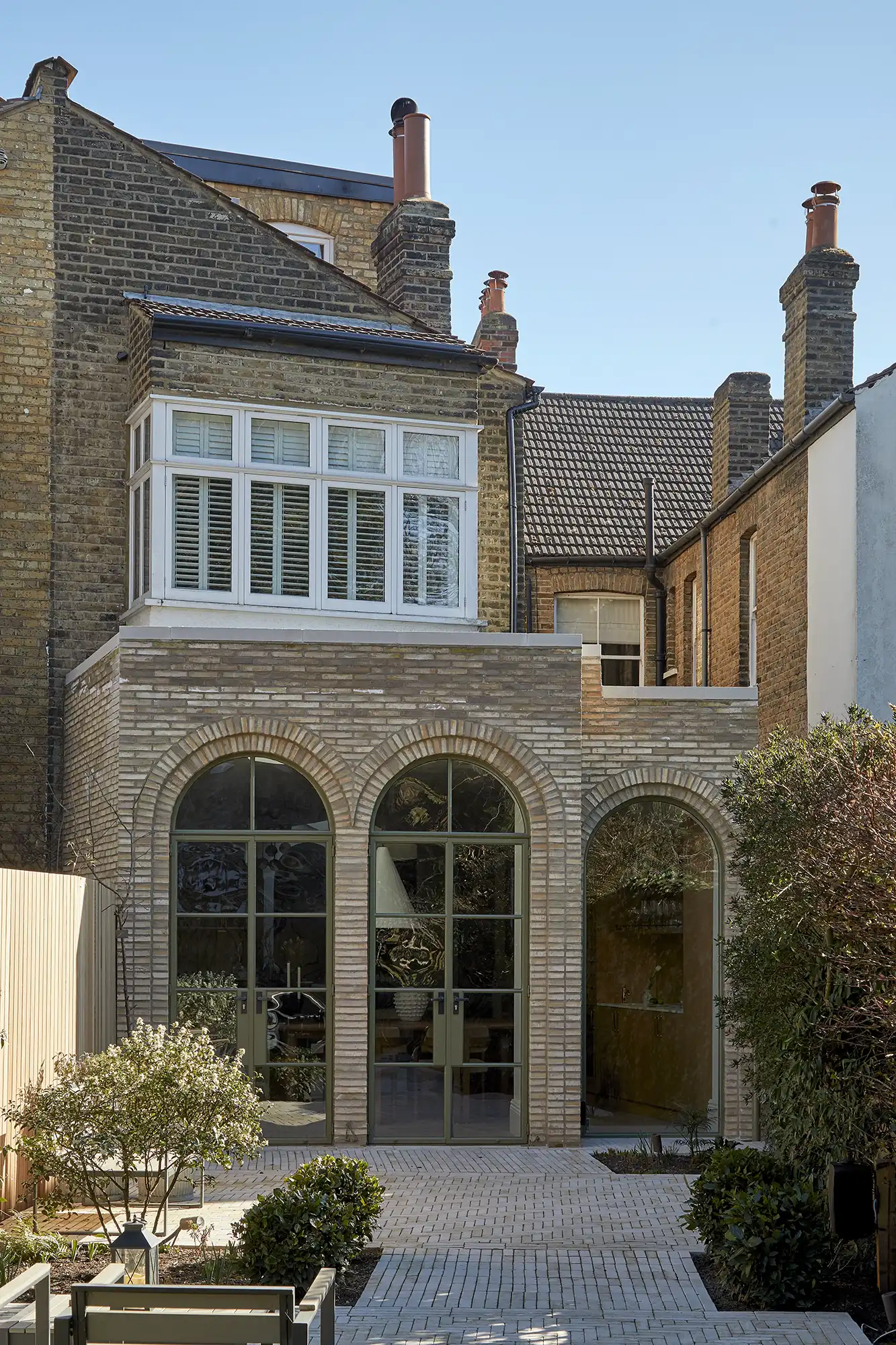 A modern brick house exterior featuring arched windows and a landscaped garden with pavers and greenery. Clear blue sky overhead.