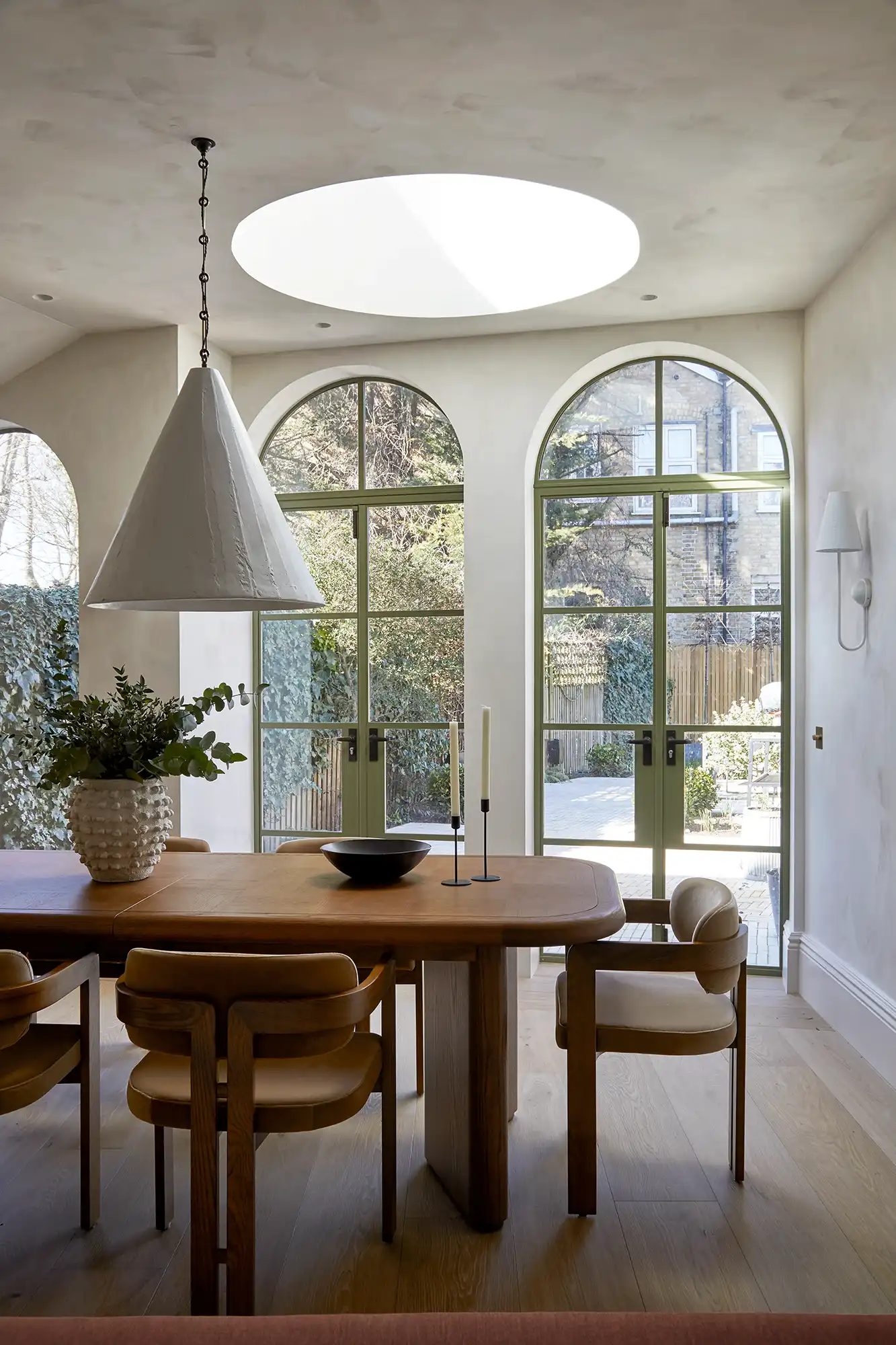 Bright dining area featuring a wooden table, light fixtures, and large arched windows, offering a view of a lush outdoor space.