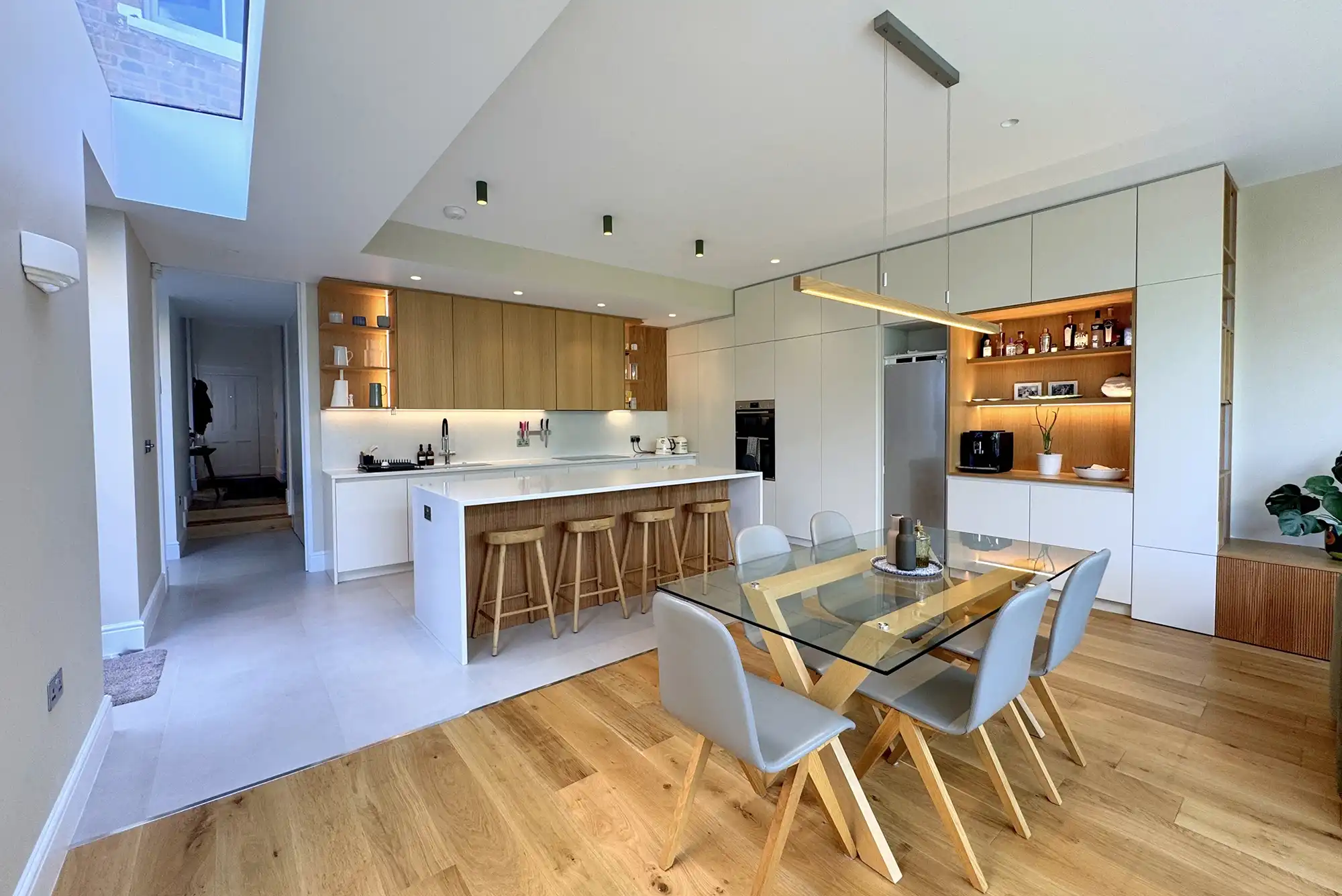 Modern kitchen and dining area with wooden elements, a glass table, and sleek appliances, featuring warm lighting and neutral tones.