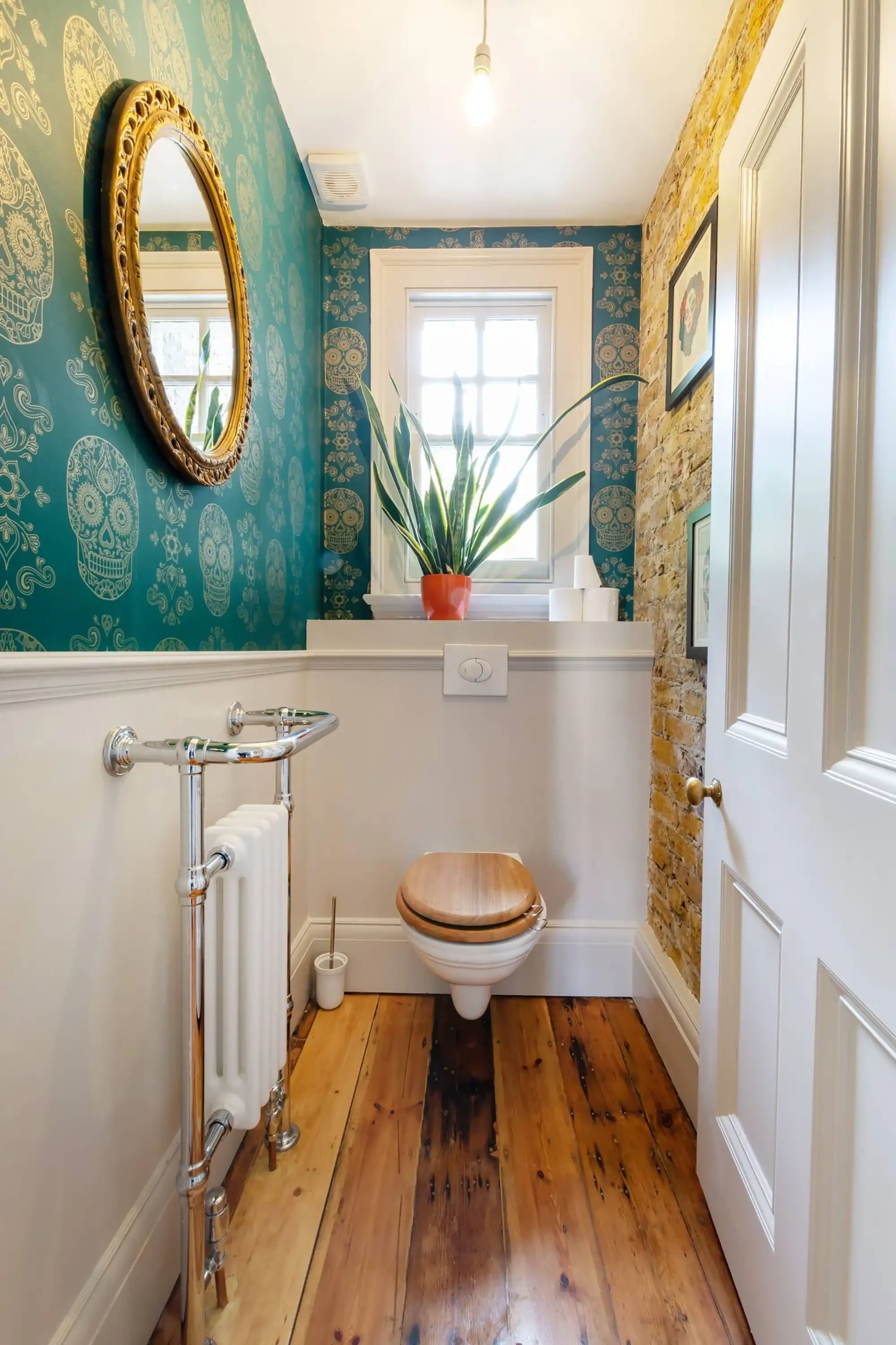 Bright bathroom featuring teal skull-patterned wallpaper, a wooden toilet seat, and a potted plant by a window.