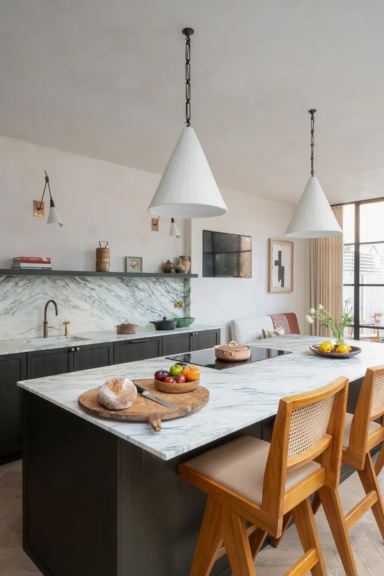 Modern kitchen with marble countertops, wooden stools, pendant lights, and decorative items. Fresh fruits on display.